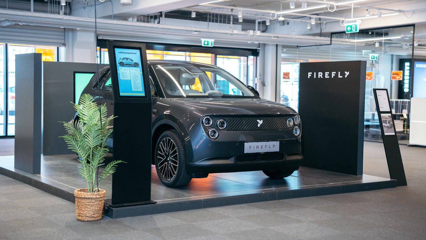 Et billede af et kontor med skriveborde og computere, set gennem vandrette persienner.