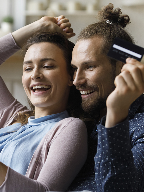 Et smilende par sidder i en sofa med en smartphone og et kreditkort i hånden og ser ud til at shoppe online sammen.